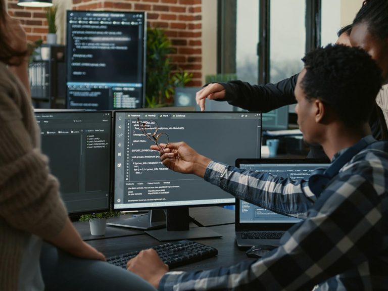 Person zeigt auf einen Computerbildschirm mit Programmiercode in einer Büroumgebung.