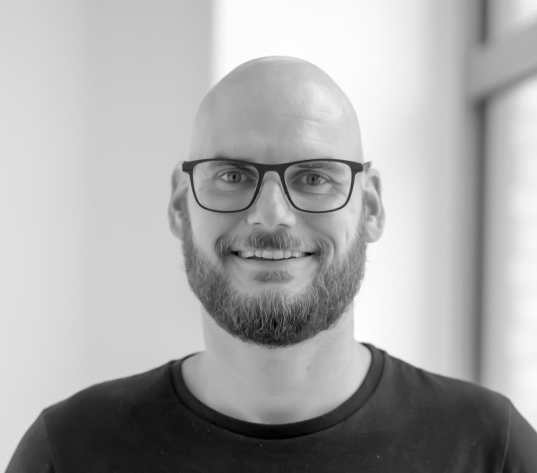 Mann mit Glatze und Brille, lächelt vor einer hellen Wand.