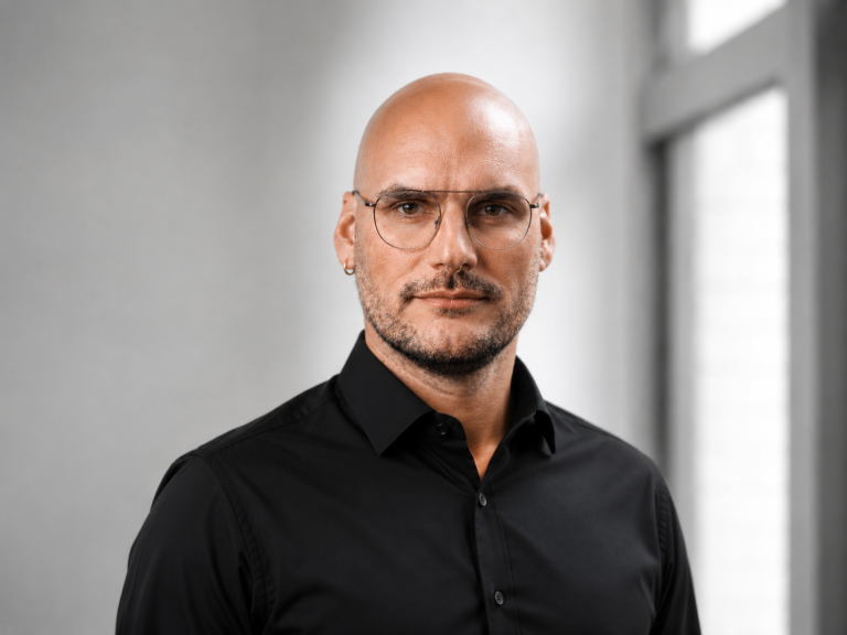 Mann mit Glatze und Brille, lächelt vor einer hellen Wand.